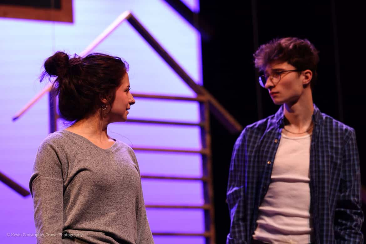 two actors, solemn, house shaped shopping shelves and light blue and purple cyc lighting are behind them